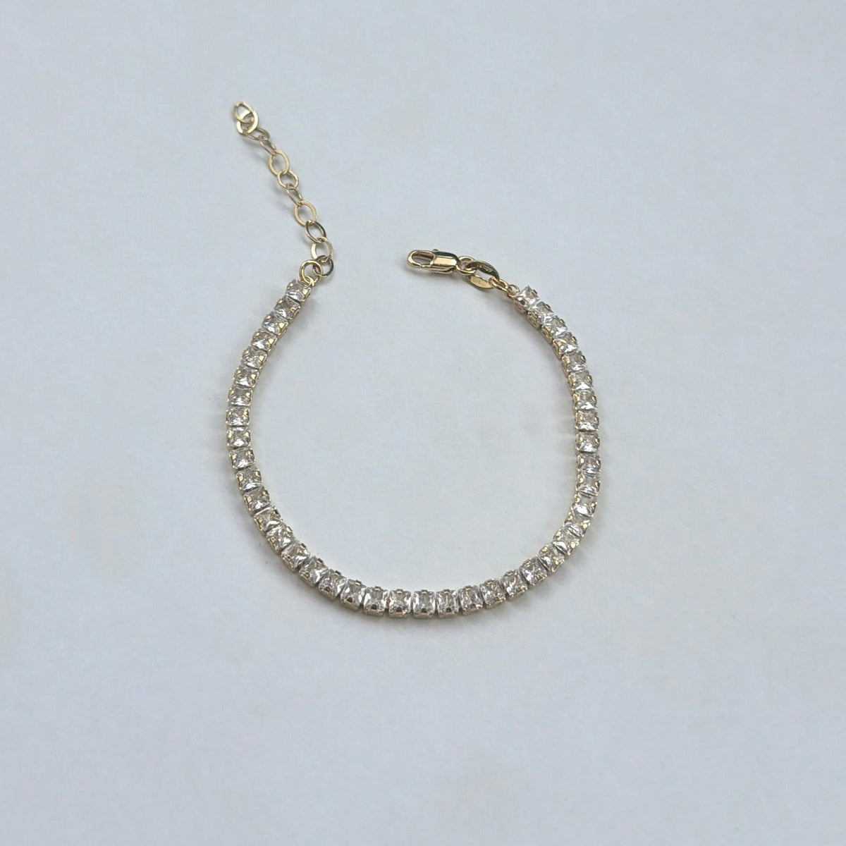 A gold tennis bracelet with clear round-cut stones, featuring an extender chain and lobster clasp, displayed on a light gray background.