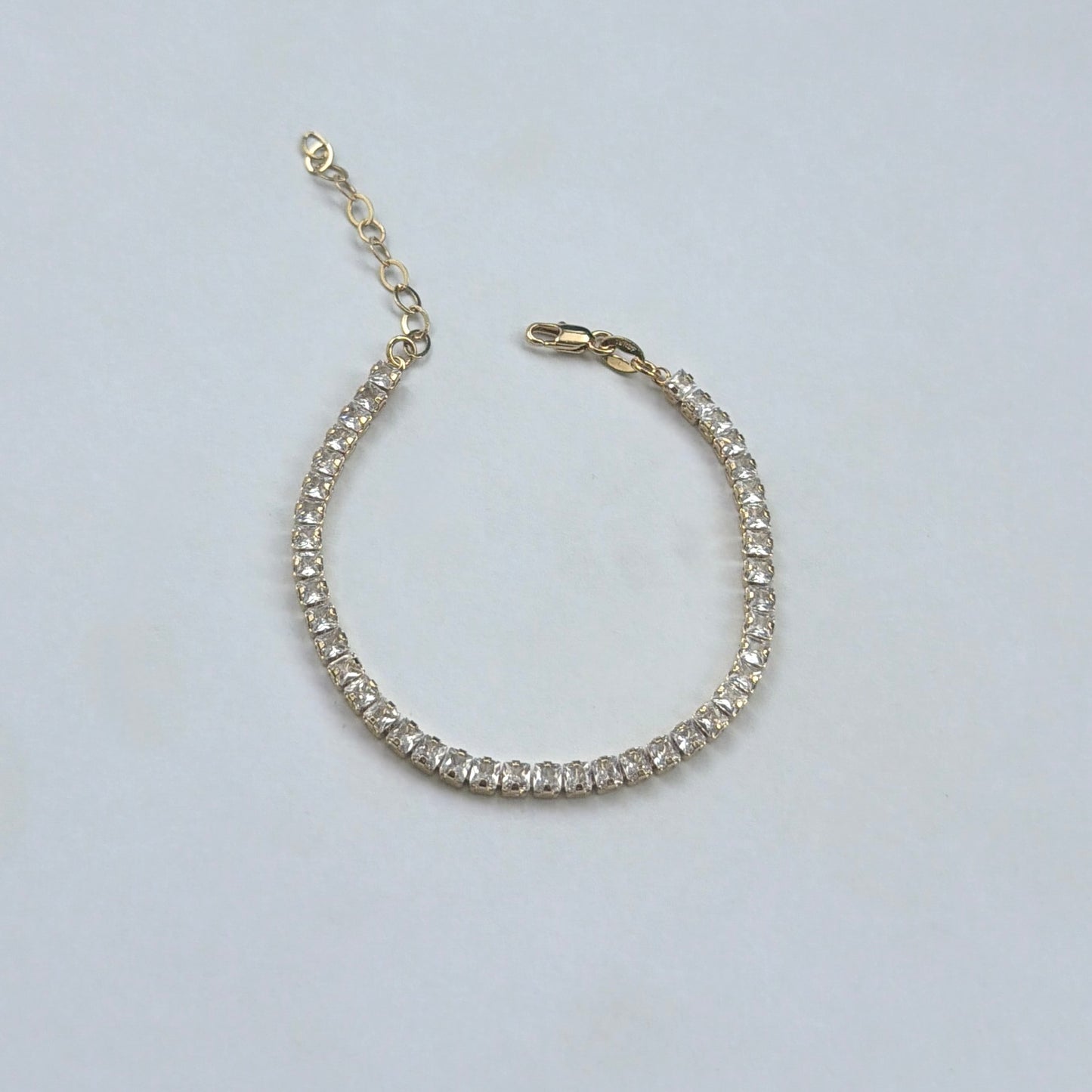 A gold tennis bracelet with clear round-cut stones, featuring an extender chain and lobster clasp, displayed on a light gray background.