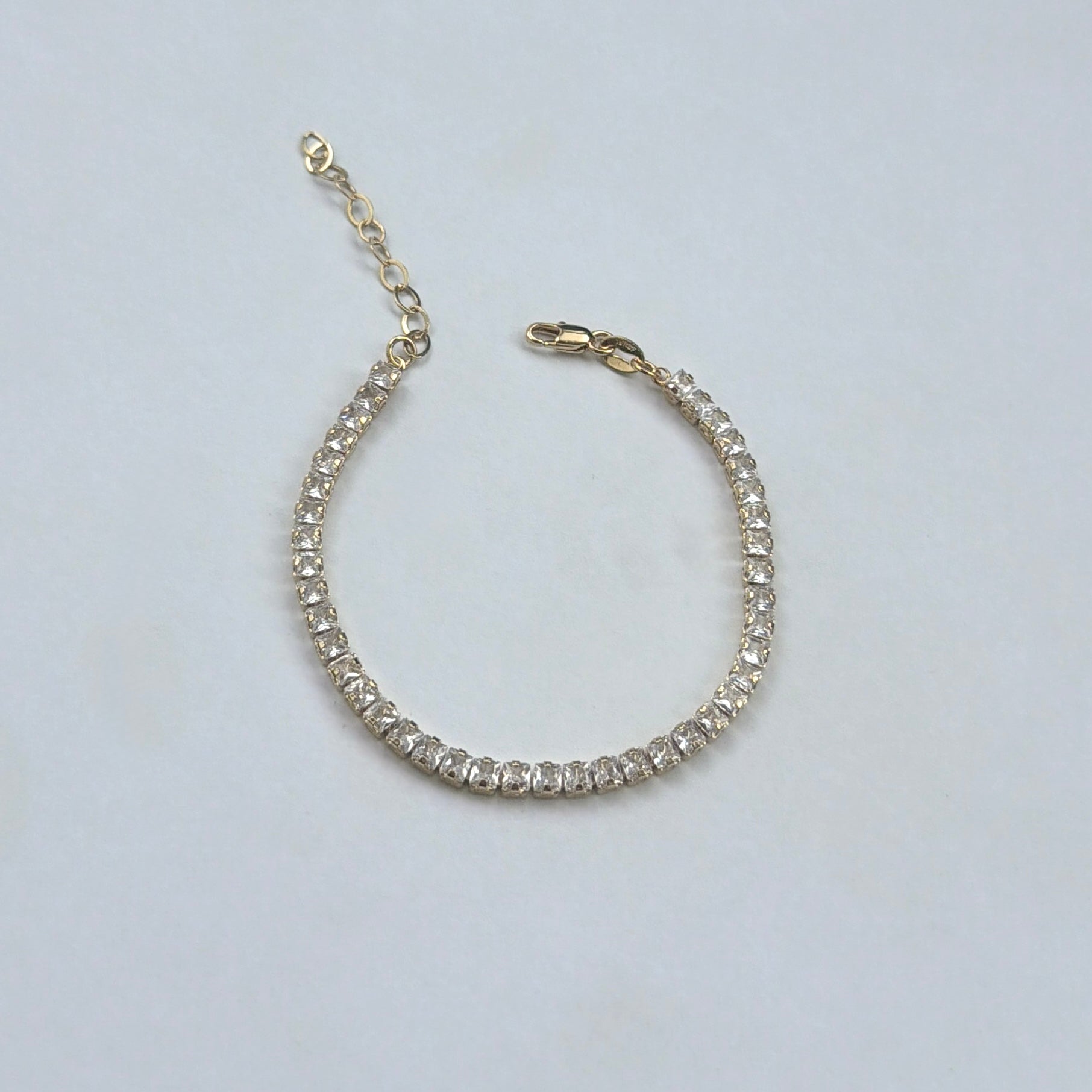 A gold tennis bracelet with clear round-cut stones, featuring an extender chain and lobster clasp, displayed on a light gray background.