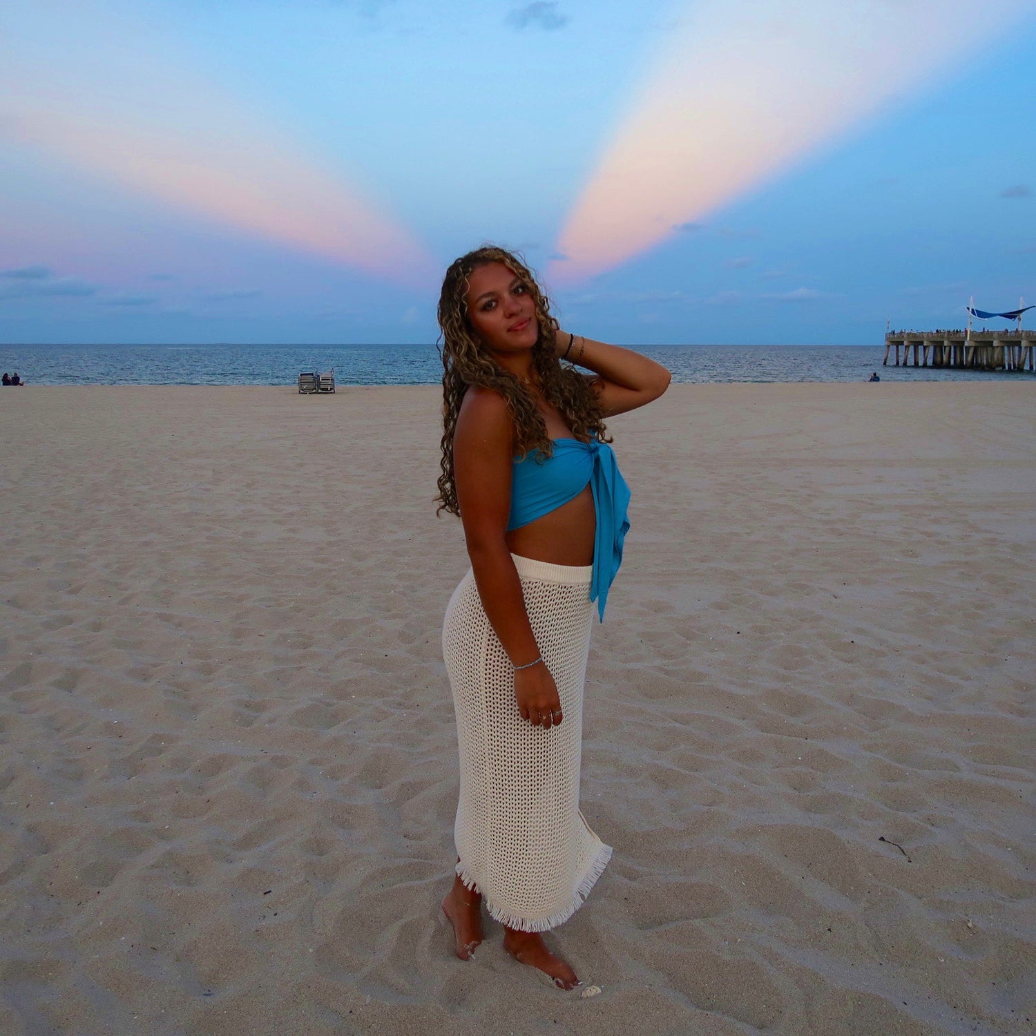 Woman in blue top and white skirt standing on a beach with a rainbow in the sky.
