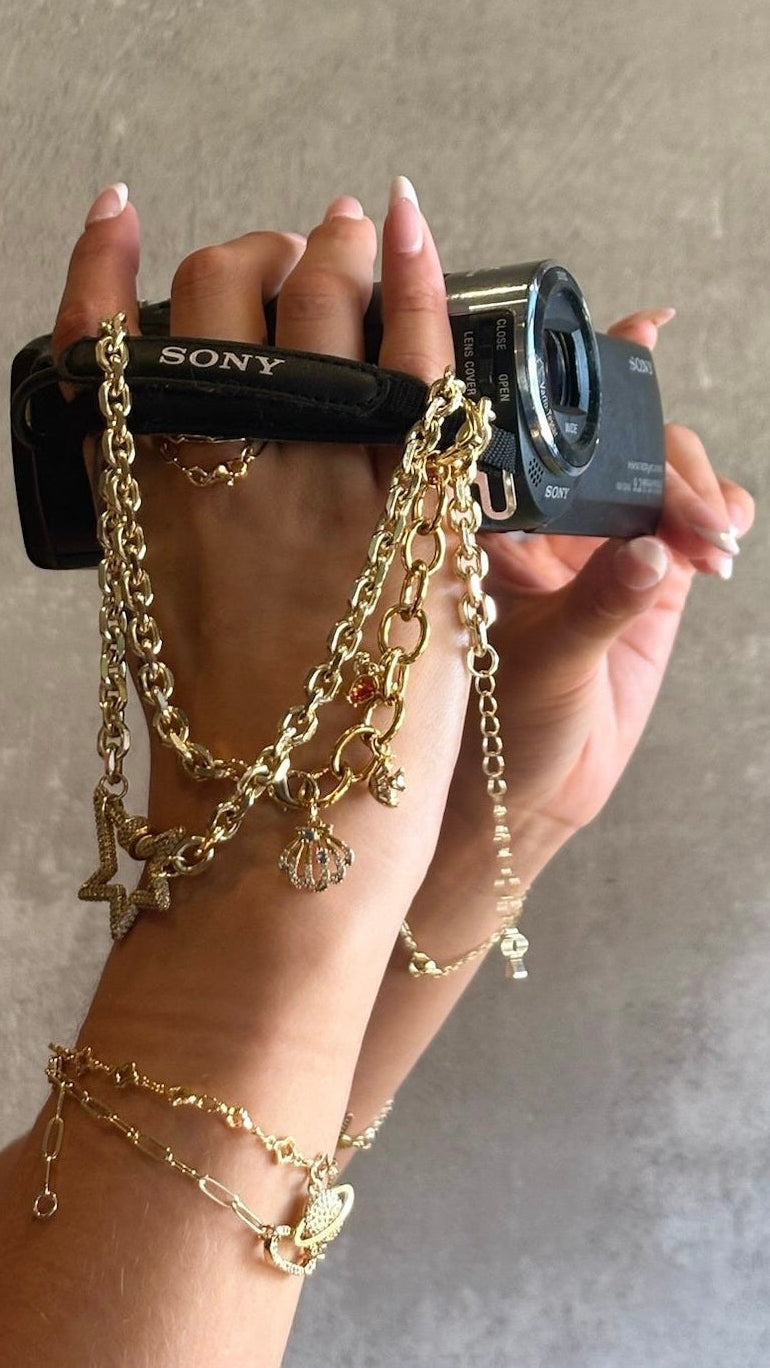 A person holds a black Sony camera with multiple draped gold chains and charms, including a small shell charm, against a textured gray concrete wall.