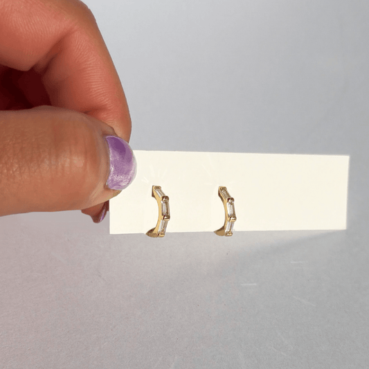 Gold earrings held by a hand over a white card with a plain background