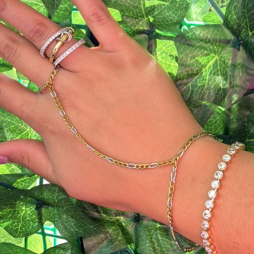 Hand wearing gold chain bracelet and ring with green leaves in the background